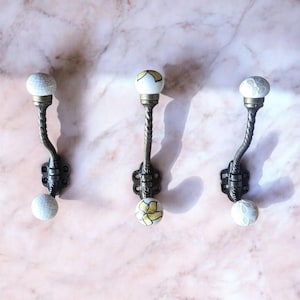 May include: Three decorative wall hooks with a vintage design. Each hook has a dark metal base and a twisted stem. The top and bottom of each hook has a white ceramic ball with a crackle glaze or floral design.