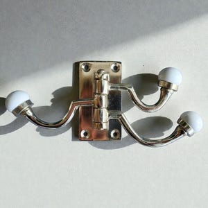 Könnte beinhalten: Ein Wandhaken aus Messing und Silber mit drei gebogenen Armen, die jeweils mit einem weißen, abgerundeten Knauf versehen sind. Der Haken ist auf einer rechteckigen Messingplatte mit Schraubenlöchern montiert.