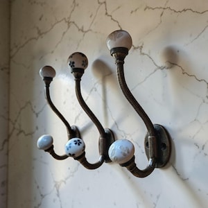 May include: Three antique-style wall hooks with a decorative design. The hooks have a dark bronze finish with a curved shape. Each hook features a white ceramic knob with a blue floral pattern. The hooks are mounted on a marble-patterned wall.
