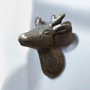 Puede incluir: Una escultura de cabeza de ciervo de color bronce con astas detalladas y una superficie texturizada. La escultura es un elemento decorativo, probablemente para montaje en pared, y tiene un aspecto rústico y antiguo. La cabeza del ciervo mira hacia la izquierda.