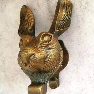 May include: A bronze-colored door knocker in the shape of a rabbit's head. The knocker features detailed fur texture and a loop for knocking. The rabbit's ears are upright, and the knocker is mounted on a wall.