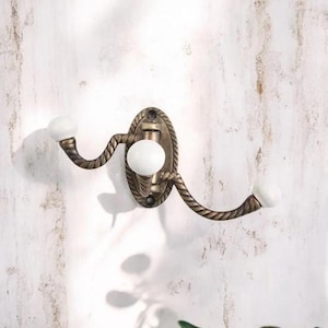 K&ouml;nnte beinhalten: Ein dekorativer Wandhaken mit Bronze-Finish und Seil-Design. Er hat drei wei&szlig;e Keramikkn&ouml;pfe. Die ovale R&uuml;ckplatte ist an einer wei&szlig;en Wand befestigt. Der Haken ist zum Aufh&auml;ngen von Gegenst&auml;nden gedacht.