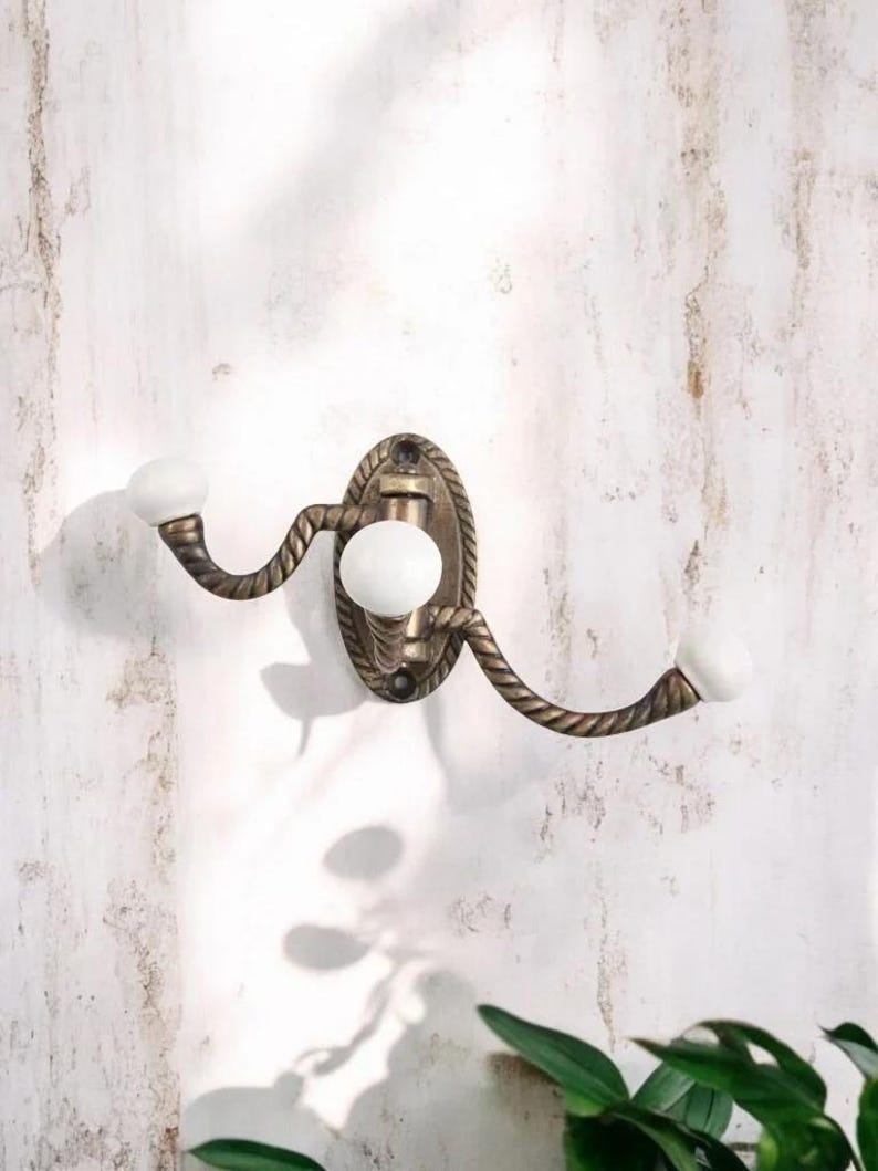 K&ouml;nnte beinhalten: Ein dekorativer Wandhaken mit Bronze-Finish und Seil-Design. Er hat drei wei&szlig;e Keramikkn&ouml;pfe. Die ovale R&uuml;ckplatte ist an einer wei&szlig;en Wand befestigt. Der Haken ist zum Aufh&auml;ngen von Gegenst&auml;nden gedacht.