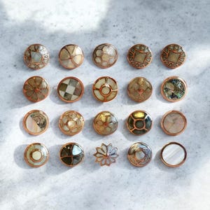 May include: An assortment of decorative buttons with copper-colored rims. The buttons feature various designs, including geometric patterns, floral motifs, and iridescent shell inlays. The buttons are arranged on a light gray surface.