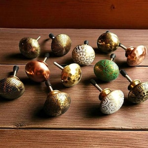 May include: A collection of decorative cabinet knobs in various metallic finishes, including gold, copper, and bronze. The knobs feature diverse textures and patterns, such as hammered, etched, and floral designs. Each knob has a black stem.