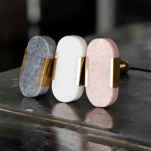 Puede incluir: Tres pomos de armario de forma ovalada con un ribete de metal dorado. Los pomos son de mármol en gris, blanco y rosa.