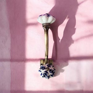 May include: A white ceramic hook with blue floral designs. The hook is attached to a gold metal base and has a curved arm.