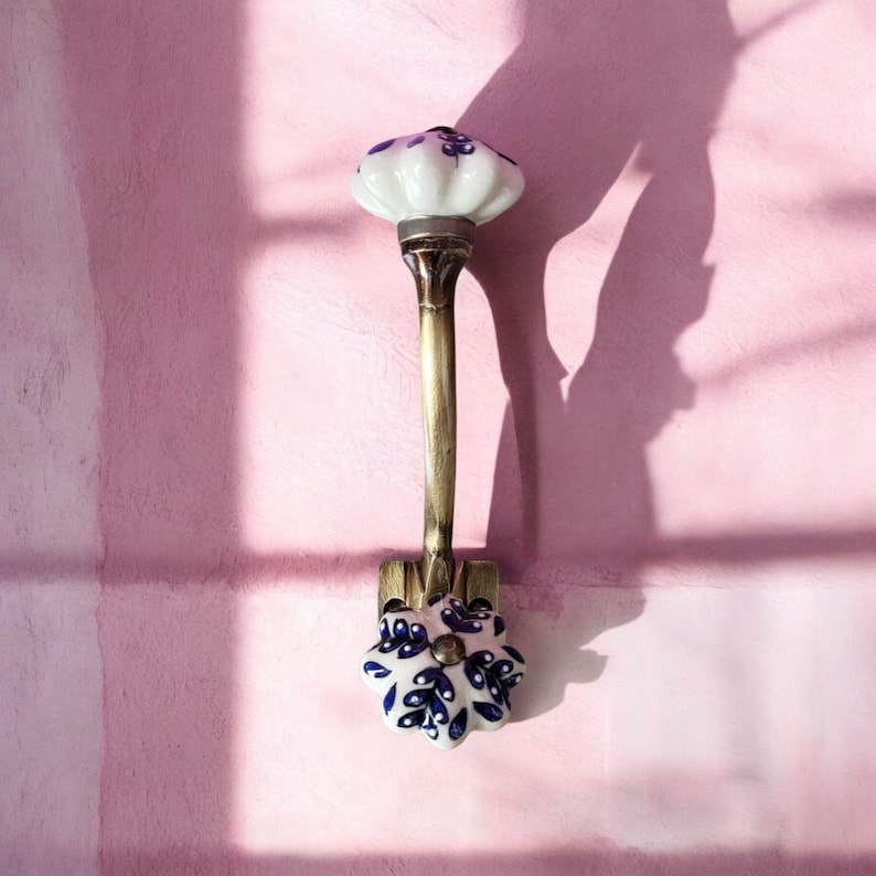 May include: A white ceramic hook with blue floral designs. The hook is attached to a gold metal base and has a curved arm.