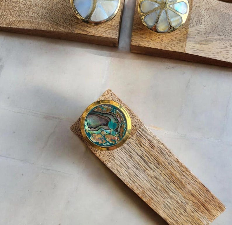 Pu&ograve; includere: Tre pomelli rotondi decorativi per armadi con base in metallo dorato e intarsi in madreperla di abalone iridescente. I pomelli sono esposti su blocchi di legno.