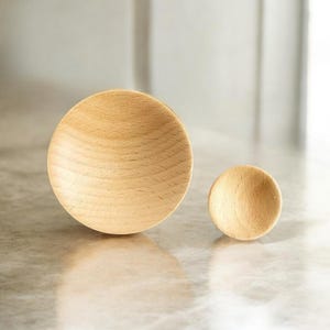 Op de afbeelding: Twee houten kommen, een grote en een kleine, worden getoond op een lichtgekleurd oppervlak. De kommen zijn gemaakt van lichtgekleurd hout en hebben een gladde, afgeronde vorm.