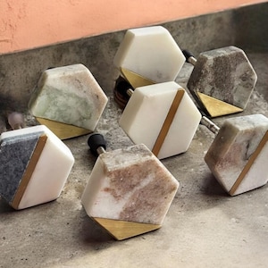 May include: Set of six hexagonal cabinet knobs with a marble and brass design. The knobs have a white marble base with a gold brass accent.