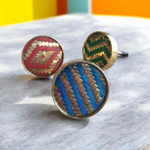 Könnte beinhalten: Drei dekorative Schrankknöpfe mit gewebten Mustern. Einer ist rot mit goldenen Akzenten, ein anderer grün mit Gold und der dritte blau mit Gold. Die Knöpfe haben ein kreisförmiges Design und eine goldfarbene Basis.