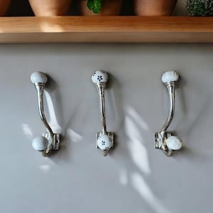 May include: Three silver hooks with white ceramic knobs featuring a black floral design. The hooks are mounted on a light grey wall.