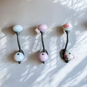 Op de afbeelding: Drie wandhaken met donker metalen beslag en keramische knoppen. De eerste haak heeft witte knoppen met roze strepen, de tweede heeft effen roze knoppen en de derde heeft witte knoppen met een roze bloemenpatroon.