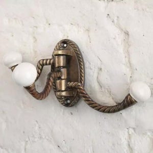 K&ouml;nnte beinhalten: Antiker Wandhaken aus Messing mit einer dekorativen ovalen R&uuml;ckplatte und zwei gebogenen Armen. Die Arme haben ein seilartiges Design und sind mit wei&szlig;en Keramikkn&ouml;pfen versehen. Der Haken ist an einer strukturierten wei&szlig;en Backsteinmauer befestigt.