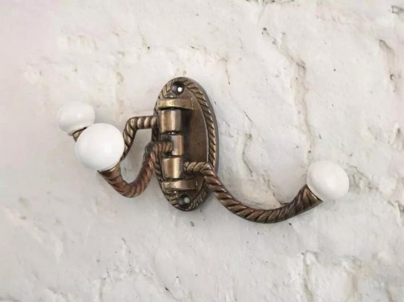 K&ouml;nnte beinhalten: Antiker Wandhaken aus Messing mit einer dekorativen ovalen R&uuml;ckplatte und zwei gebogenen Armen. Die Arme haben ein seilartiges Design und sind mit wei&szlig;en Keramikkn&ouml;pfen versehen. Der Haken ist an einer strukturierten wei&szlig;en Backsteinmauer befestigt.