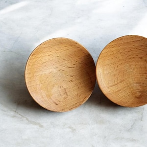 May include: Two small, round wooden bowls. The bowls are made of light brown wood and have a smooth, polished finish.
