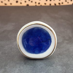 K&ouml;nnte beinhalten: Ein wei&szlig;er Keramik-Knopf mit goldfarbenem Rand und einem blauen Zentrum. Das blaue Zentrum hat einen craquel&eacute;-Effekt.