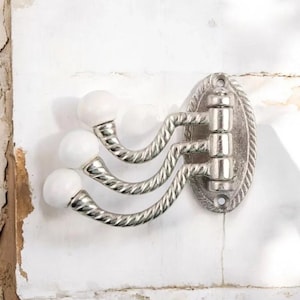 K&ouml;nnte beinhalten: Ein dekorativer Wandhaken mit drei gebogenen Armen und wei&szlig;en, kugelf&ouml;rmigen Enden. Die Metallhalterung hat eine silberne Oberfl&auml;che und eine ovale R&uuml;ckplatte mit einem seilartigen Rand. Der Haken ist an einer wei&szlig;en Wand befestigt.