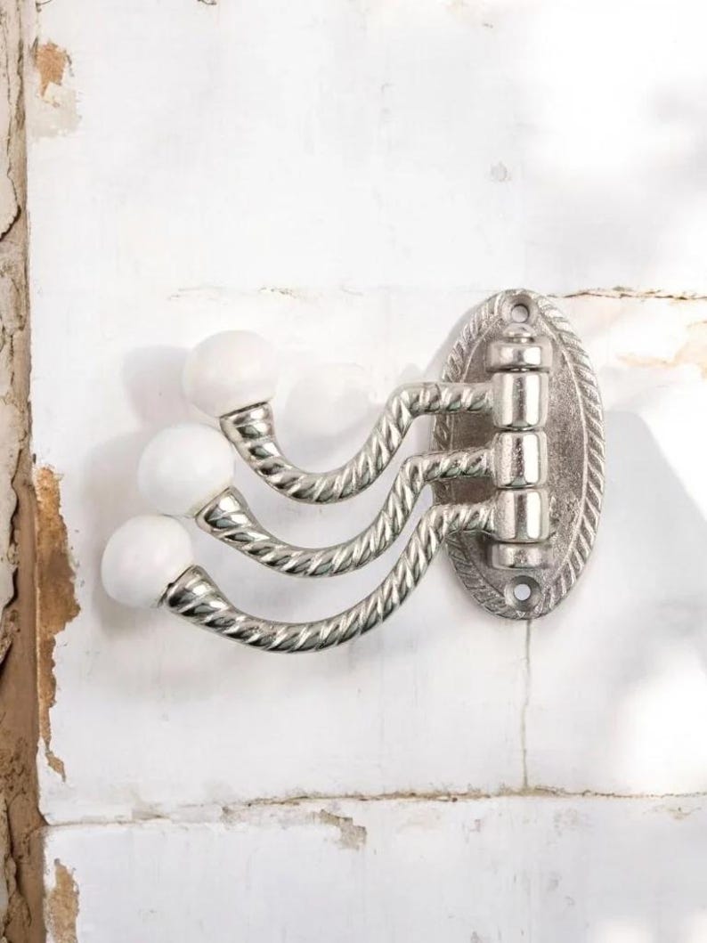 K&ouml;nnte beinhalten: Ein dekorativer Wandhaken mit drei gebogenen Armen und wei&szlig;en, kugelf&ouml;rmigen Enden. Die Metallhalterung hat eine silberne Oberfl&auml;che und eine ovale R&uuml;ckplatte mit einem seilartigen Rand. Der Haken ist an einer wei&szlig;en Wand befestigt.