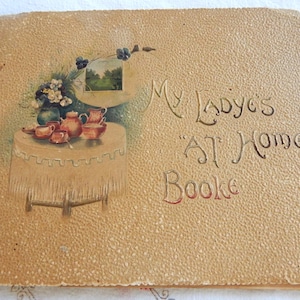 May include: Vintage book cover with embossed texture and the title "My Lady's 'At Home' Booke." The cover features a painted illustration of a table set with a tea service and a small landscape painting. The color palette is primarily beige and brown.