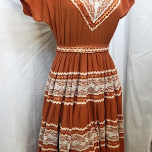May include: A rust-colored dress featuring short sleeves, a V-neckline, and a cinched waist. The garment is decorated with horizontal bands of white floral lace trim and metallic copper-toned ribbon accents throughout the bodice and skirt.