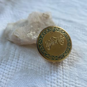 May include: A round, gold-toned compact with the text "Djer Kiss" in elegant script. The compact has a green floral border and is set against a white, textured background. The compact appears to be vintage.