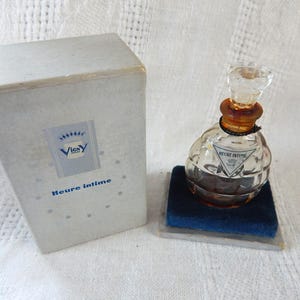 May include: Vintage perfume bottle with a clear, textured glass body and a clear stopper. The bottle sits on a blue velvet pad next to a light blue box with the text "Vicy Heure intime".