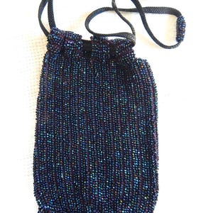May include: A small drawstring pouch covered in iridescent dark blue and purple beads. The bag features a vertical striped pattern of shimmering beads, a black cord drawstring closure, and a fringe of matching beaded strands at the bottom edge.