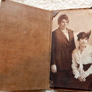 May include: A vintage photo album with a sepia-toned photo of two women. The women are wearing dark clothing and hats. The photo is titled "Greetings from New York".