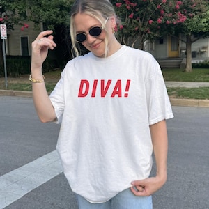May include: White t-shirt with the word "DIVA!" printed in red. The shirt is a casual style, suitable for everyday wear. The text is bold and centered on the front of the shirt.