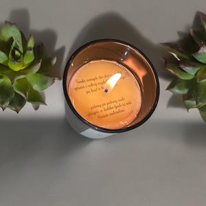 May include: A small, round, amber-colored candle with a white label. The label has text in a brown font that reads "Svaki osmijch što danas spremi i sakrij najdet kad ti be pa jednog po jednog radi prisjeti se koliko ljudi te voli Sretan rodendan!"