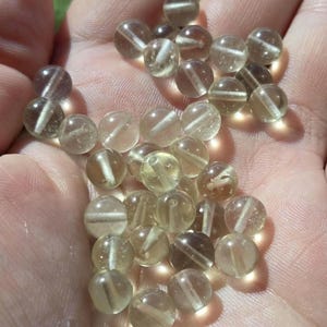 Puede incluir: Una colección de pequeñas cuentas redondas y transparentes con un tono amarillo-marrón claro. Cada cuenta tiene una línea recta blanca que atraviesa su centro. Las cuentas se sostienen en una mano, iluminadas por la luz del sol.