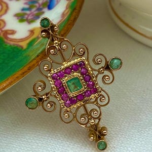 Puede incluir: Broche dorado con un cuadrado central de piedras preciosas rosas, rodeado de gemas rosas más pequeñas y una gema cuadrada verde en el centro. Las gemas verdes están engastadas en los extremos del diseño en forma de cruz del broche. El broche tiene un diseño de filigrana ornamentado.
