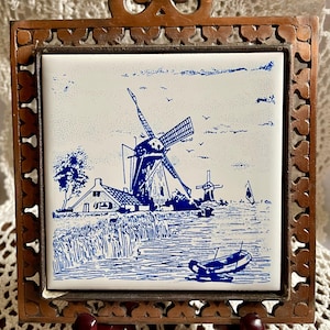 May include: A square ceramic tile with a blue and white windmill scene, set in a decorative wooden frame. The tile depicts a rural landscape with windmills, a boat, and a house. The frame has an ornate, openwork design.