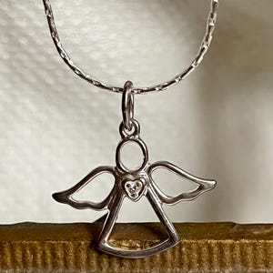 K&ouml;nnte beinhalten: Ein silberner Engelsanh&auml;nger mit herzf&ouml;rmigem Mittelteil, besetzt mit einem kleinen, klaren Stein, h&auml;ngt an einer zarten Silberkette. Der Engel hat ausgebreitete Fl&uuml;gel und einen Heiligenschein. Der Schmuck wird vor neutralem Hintergrund pr&auml;sentiert.