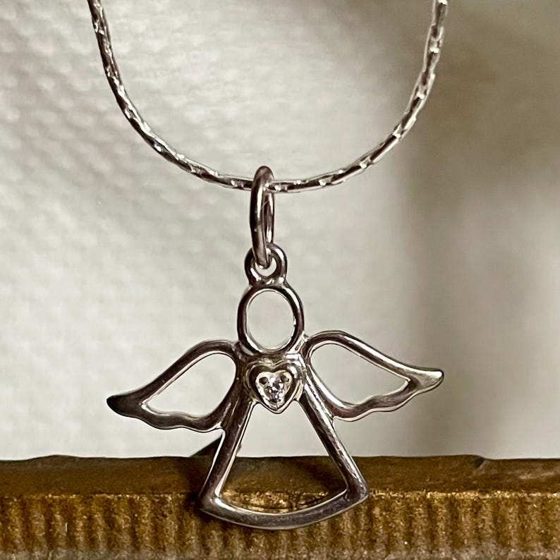 K&ouml;nnte beinhalten: Ein silberner Engelsanh&auml;nger mit herzf&ouml;rmigem Mittelteil, besetzt mit einem kleinen, klaren Stein, h&auml;ngt an einer zarten Silberkette. Der Engel hat ausgebreitete Fl&uuml;gel und einen Heiligenschein. Der Schmuck wird vor neutralem Hintergrund pr&auml;sentiert.