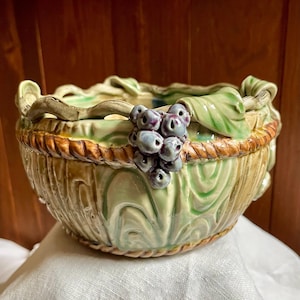 May include: A ceramic bowl featuring a light green glazed finish with a textured surface. The rim is decorated with a sculpted rope pattern and three-dimensional clusters of purple grapes and green leaves attached to the exterior side.