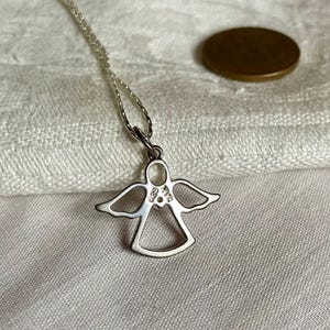 K&ouml;nnte beinhalten: Eine silberne Engelsanh&auml;nger-Halskette. Der Anh&auml;nger ist eine stilisierte Umrisszeichnung eines Engels mit ausgebreiteten Fl&uuml;geln. Die Halskette ist filigran. Im Hintergrund ist eine M&uuml;nze zu sehen.