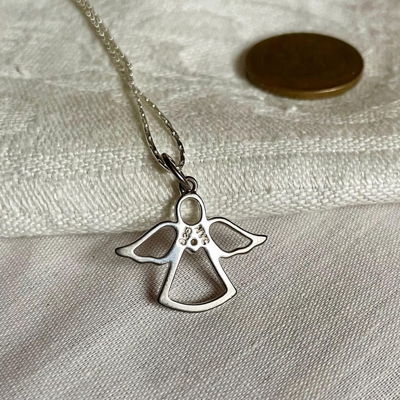 K&ouml;nnte beinhalten: Eine silberne Engelsanh&auml;nger-Halskette. Der Anh&auml;nger ist eine stilisierte Umrisszeichnung eines Engels mit ausgebreiteten Fl&uuml;geln. Die Halskette ist filigran. Im Hintergrund ist eine M&uuml;nze zu sehen.