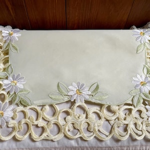 May include: A rectangular, pale yellow table runner with embroidered daisy flowers and green leaves. The edges feature a scalloped, openwork design in a soft yellow color. The daisies have white petals and yellow centers.