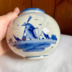 May include: A small, round ceramic vase with a blue and white windmill design. The vase has a blue rim and the word "Holland" is written in blue. The design includes a windmill, houses, and birds.