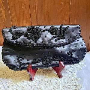 May include: A black satin clutch bag featuring intricate black beadwork in floral and leaf patterns. The exterior is adorned with scattered black beads across the surface, and the flap closure is decorated with larger, detailed beaded flower motifs.