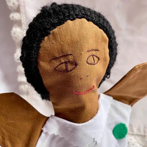 May include: Handmade cloth doll with brown skin, black yarn hair, and embroidered facial features. The doll wears a white dress with a green circle accent. The doll's arms are outstretched, giving it a welcoming appearance.
