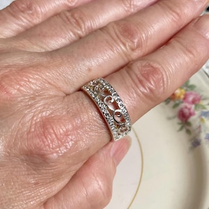 May include: Silver-toned ring with an openwork filigree design. The ring is encrusted with small, clear gemstones along the top and sides. The ring is on a finger, showcasing its intricate details and sparkling appearance.