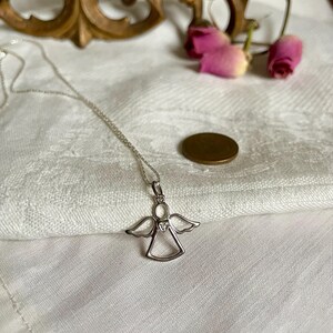 K&ouml;nnte beinhalten: Eine silberne Engelsanh&auml;nger-Halskette mit zarter Kette. Der Engelsanh&auml;nger hat ausgebreitete Fl&uuml;gel und ein herzf&ouml;rmiges Detail. Die Halskette liegt auf einer wei&szlig;en, strukturierten Oberfl&auml;che, mit einer M&uuml;nze und getrockneten Rosen im Hintergrund.