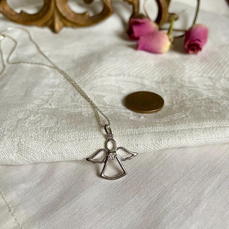 K&ouml;nnte beinhalten: Eine silberne Engelsanh&auml;nger-Halskette mit zarter Kette. Der Engelsanh&auml;nger hat ausgebreitete Fl&uuml;gel und ein herzf&ouml;rmiges Detail. Die Halskette liegt auf einer wei&szlig;en, strukturierten Oberfl&auml;che, mit einer M&uuml;nze und getrockneten Rosen im Hintergrund.