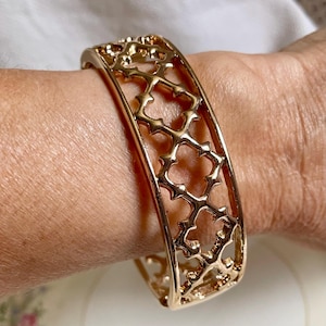 May include: A gold-colored cuff bracelet with an openwork design. The bracelet features a repeating pattern of stylized floral or geometric shapes. The bracelet is worn on a wrist and appears to be made of metal.