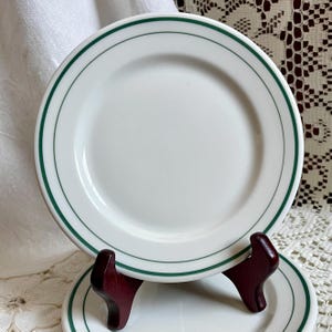 May include: White ceramic plate with a green double-line border. The plate is round and sits on a dark wooden plate stand. The plate is clean and simple, suitable for everyday use.