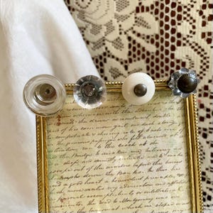 May include: Four decorative cabinet knobs in a row. The knobs are made of glass, ceramic, and metal. The first knob is clear glass, the second is a clear faceted glass, the third is white ceramic, and the fourth is a silver metal flower shape.